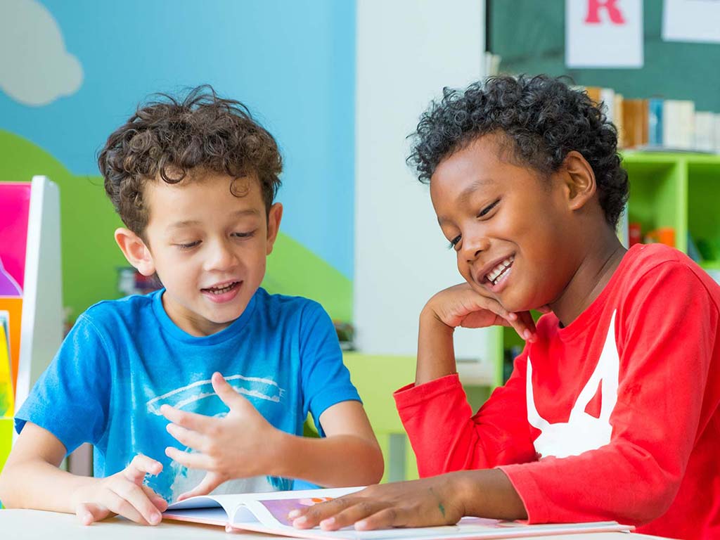 Two young boys sit at a table, reading and discussing a book together in a brightly colored classroom.