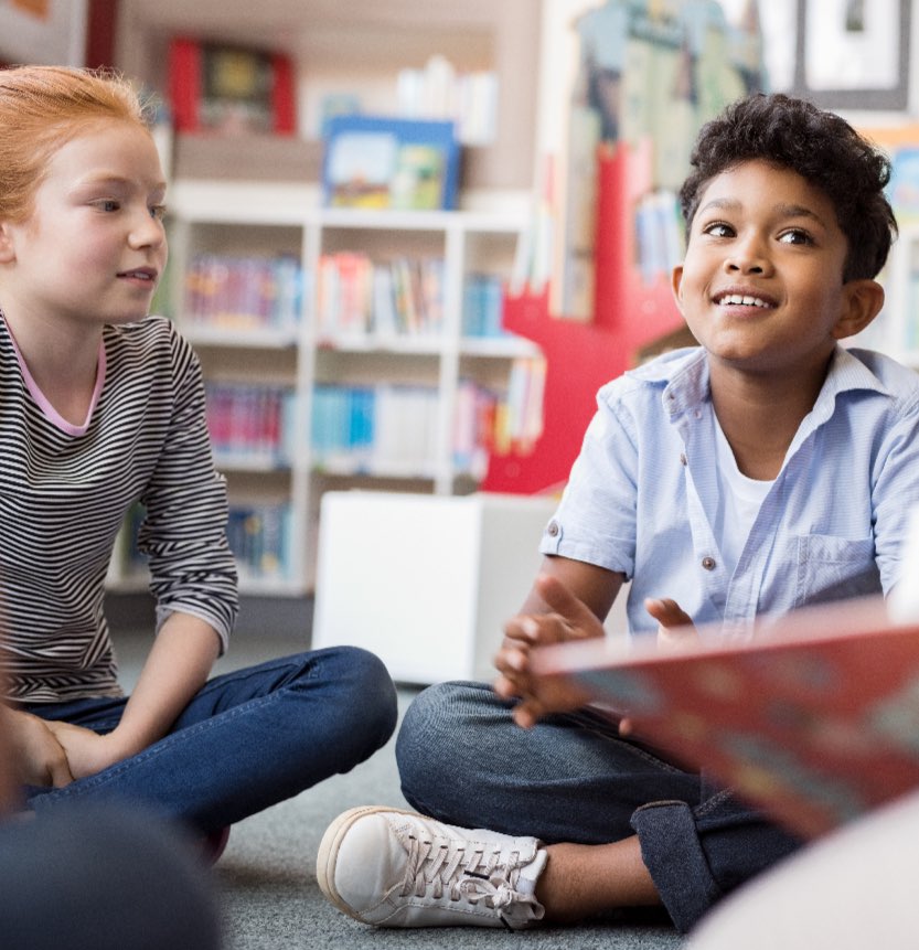 Two children sit cross-legged on the floor in a library, talking and smiling, with shelves of books visible in the background.